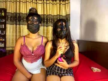 Media: Video of two women in lingerie, one in a black mask, sitting on a red bed, with a beige curtain in the background.