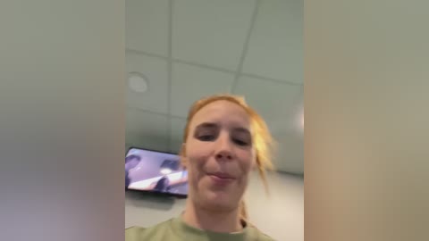 Media: Video of a smiling, fair-skinned woman with red hair, wearing a green top, standing in a modern office with a TV screen displaying a person on the left wall.