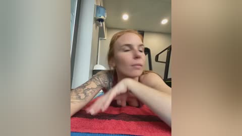 Media: A video shows a blonde woman with a tattoo sleeve lying on a red and black striped towel in a gym, eyes closed, with a blurred background.