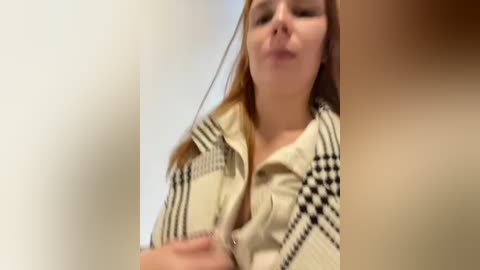 Media: A close-up video of a young woman with long, straight red hair, wearing a beige and black checkered shirt. She has fair skin and is slightly out of focus, creating a soft blur around her.