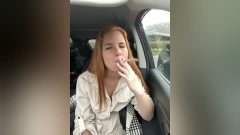 Media: Video of a red-haired woman with fair skin, wearing a beige jacket, sitting in a car, smoking a cigarette with a relaxed expression. The background shows a blurred, green landscape.