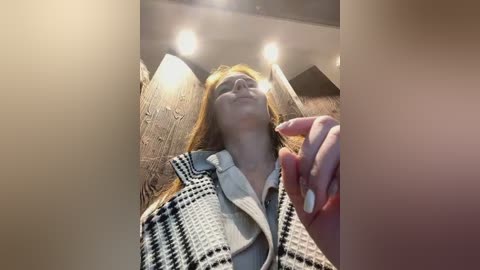 Media: Video of a young Caucasian woman with long, straight auburn hair, wearing a patterned black-and-white blazer over a light-colored shirt. She stands in a dimly lit, textured stone-walled room, with a hand raised, fingers extended.