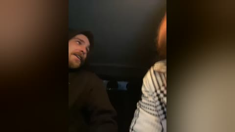 Media: A dimly lit video captures a man with a beard, wearing a brown coat, seated in a car, speaking to a person with a plaid shirt, partially obscured by the frame.