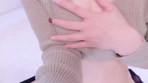 Media: Video of a person with pale skin, wearing a beige knit sweater, with hands gently touching their chest, nails painted in a dark color, white background.