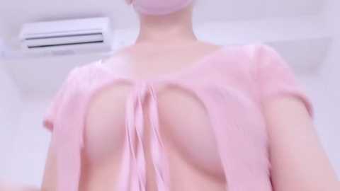 Media: Video of a pale-skinned person wearing a light pink, see-through blouse, revealing their bare breasts, with a pink face mask and an air conditioner visible in the background.