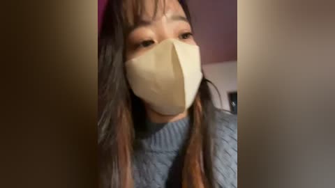 Media: A video of an Asian woman with long, straight, dark hair, wearing a beige face mask and a light blue sweater, standing against a blurred, neutral-colored background.