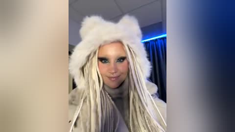 Media: Video of a blonde woman with long, white dreadlocks, wearing a white fur hat, smiling, standing in a dimly lit room with blue lighting.