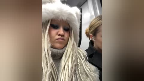 Media: Video of a blonde woman with long, braided hair, wearing a fur-trimmed hat and gray turtleneck sweater, looking pensive. A blonde woman in a dark coat is partially visible behind her.