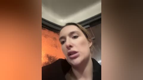 Media: A video of a woman with light skin, blonde hair, and a black blazer, standing indoors with an orange-tinted window behind her, looking slightly confused or surprised.