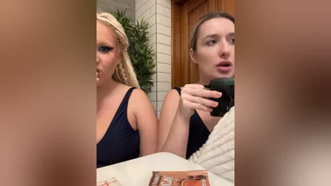 Media: Video of two women, one with blonde braids and dark makeup, the other with brown hair and natural makeup, both wearing black tops, in a bathroom, discussing something with a microphone.