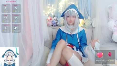 Media: Video of a young woman in a blue and white shark-themed cosplay, sitting on a couch, surrounded by soft, pastel-colored decor, and a virtual overlay displaying a character's stats.