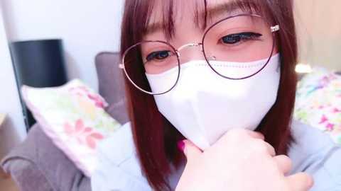 Media: Video of an East Asian woman with glasses and a white mask, holding a phone, sitting on a bed with floral pillows.