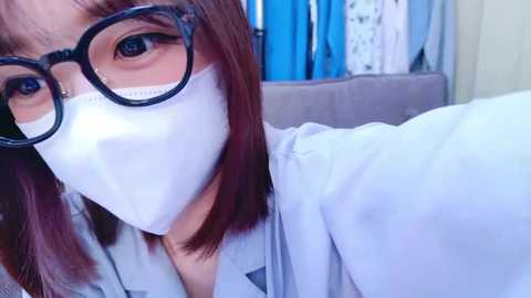 Media: Video of an East Asian woman with shoulder-length, straight brown hair, wearing black-rimmed glasses, white surgical mask, and a light-colored lab coat, in a clinical setting with blue curtains and medical equipment in the background.