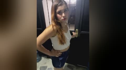 Media: Video of a young woman with long, wavy brown hair, wearing a white crop top and dark blue shorts, taking a selfie in a dimly lit room with a dark door and carpeted floor.