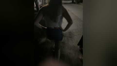 Media: A dimly lit video of a topless woman with long hair, wearing blue shorts, standing in a dark, narrow hallway, with a blurred figure partially visible in the foreground.