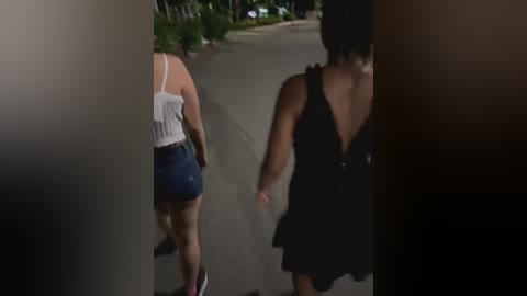 Media: Video of two women walking away from the camera. One wears a white tank top and blue shorts, the other a black dress. They are on a suburban street with trees and a sidewalk.