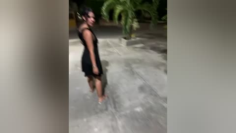 Media: Video of a dark-skinned woman in a black dress walking on a concrete path in a lush, green garden setting. Her hair is styled in a bun.