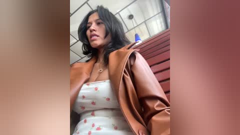 Media: Video of a woman with dark hair and medium skin tone, wearing a white floral dress and brown leather jacket, standing in a modern, brightly lit room with white ceiling tiles and red walls.