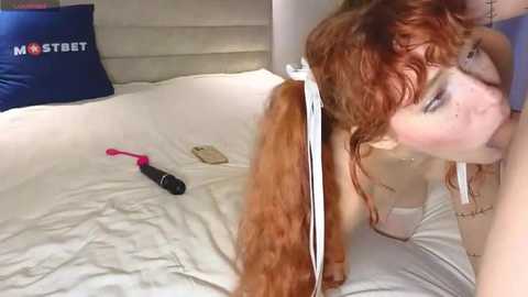 Media: Video of a fair-skinned woman with long, curly red hair, performing oral sex on a man, lying on a white bed. A pink vibrator and phone lie beside her.