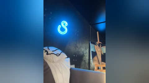 Media: Video of a dimly lit, modern bedroom with a neon blue \"S\" sign on a dark wall, a partially visible white bed with black trim, and a person in a mirror, wearing a white tank top.