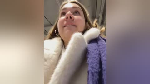 Media: Video of a young woman with light skin, brown hair, and a purple bathrobe, looking up with a neutral expression in a bathroom with white tiles and a towel hanging on the wall.