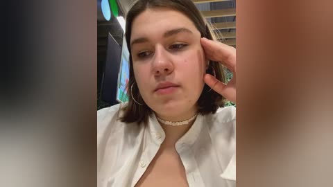 Media: A video of a young woman with fair skin and medium-length brown hair, wearing a white button-up shirt and a choker necklace, looking down with a hand on her ear in an indoor setting.