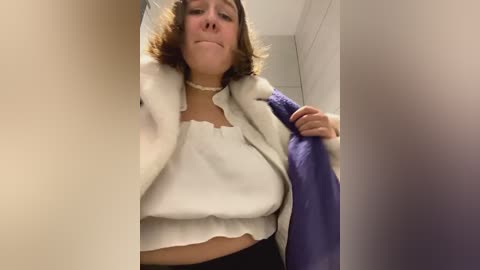 Media: Video of a woman with shoulder-length brown hair, wearing a white blouse and purple robe, holding a towel in a bathroom with white tiles.