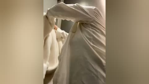 Media: Video of a person in a white robe, partially obscured, standing in a narrow hallway with beige walls, leaning against a wall with their left arm.