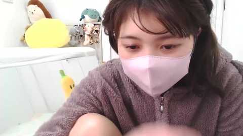 Media: A video of an Asian woman with shoulder-length dark hair, wearing a pink face mask, brown robe, and sitting in a white room with stuffed animals.