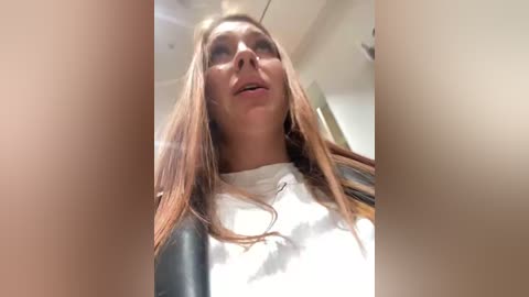 Media: A video of a young woman with long, straight, light brown hair, wearing a white shirt, captured from a low angle. The background shows an indoor setting with white walls and a ceiling.