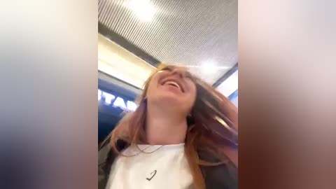 Media: Video of a laughing young woman with long, light brown hair, wearing a white tank top and black jacket, inside a modern, brightly lit space with a grid ceiling and blurred background.