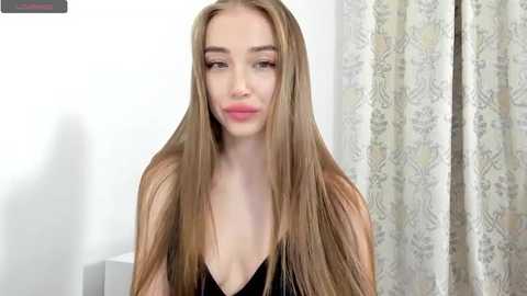 Media: Video of a fair-skinned, young woman with long, straight, light brown hair, wearing a black, low-cut top, against a white wall and patterned curtain background.