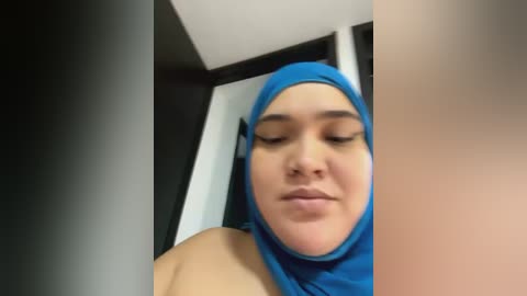 Media: Video of a light-skinned woman with a round face, wearing a blue hijab, looking down. She has a neutral expression. The background features a modern, minimalist room with white and black walls.