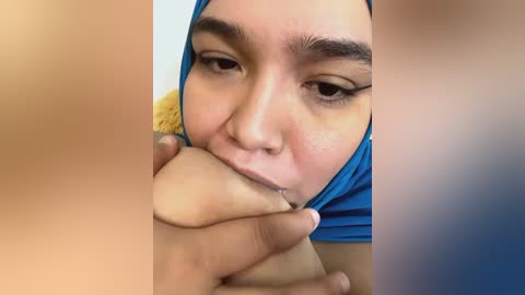 Media: Video of an Asian woman with fair skin and brown eyes, wearing a blue hijab, seductively licking a man's erect penis. The background is blurred.