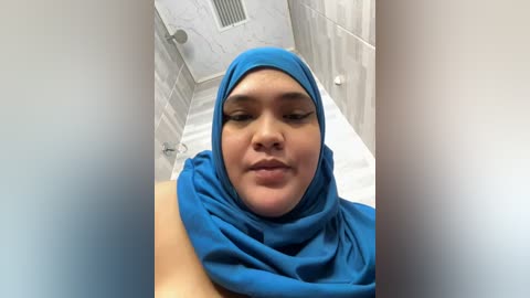 Media: Video of a young woman with medium brown skin, wearing a bright blue hijab and a white shirt, standing in a white-tiled bathroom.