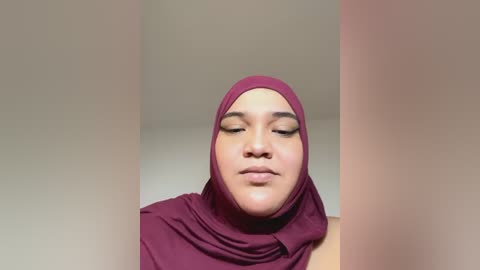 Media: Video of a serene woman with medium-brown skin, wearing a maroon hijab and matching top, eyes closed, set against a soft beige background.