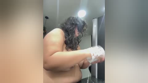 Media: Video of a nude, overweight woman with curly hair, wearing only white socks, scrubbing a mirror in a dimly lit bathroom with a small light above.