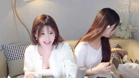 Media: Video of two Asian women with long, straight hair, smiling, sitting on a gray couch with black and white checkered pillows. One wears a white top, the other a white dress, in a modern living room.