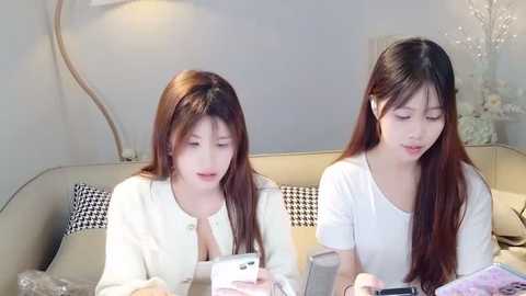 Media: Video of two East Asian women with long brown hair, wearing white shirts, sitting side-by-side at a table in a modern, minimalist room.