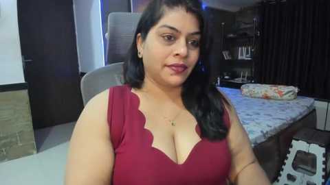 Media: Video of a plus-sized South Asian woman with medium skin tone and long black hair, wearing a deep red, scalloped neckline dress. She's indoors in a modern room with a bed, shelves, and dark curtains in the background.