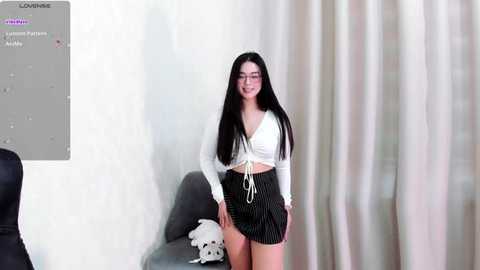 Media: Video of a young woman with long black hair, fair skin, and glasses, wearing a white crop top and high-waisted pinstripe shorts, standing in a minimalist room with beige curtains and a dark chair.