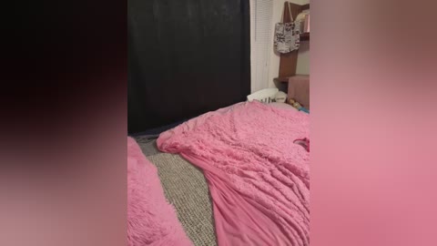 Media: Video of a pink, fluffy blanket draped over a beige bedspread, partially covering a black curtain and a white wall with a shelf holding bags and boxes.