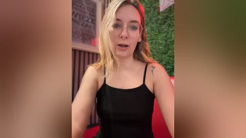 Media: Video of a fair-skinned, blonde woman with glasses and a black tank top, looking surprised. Background features a brick wall, greenery, and red furniture.