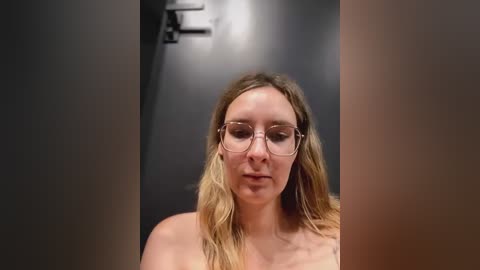 Media: Video of a nude, fair-skinned woman with long, wavy, blonde hair and thin-rimmed glasses, standing in a dimly lit, grey-toned room with a black wall and a small, out-of-focus light fixture in the background.