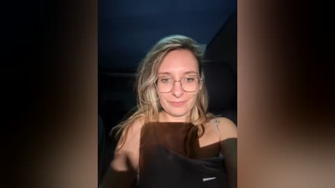 Media: Video of a young woman with long blonde hair, wearing glasses, and a black top, indoors, dim lighting, slightly blurry background.