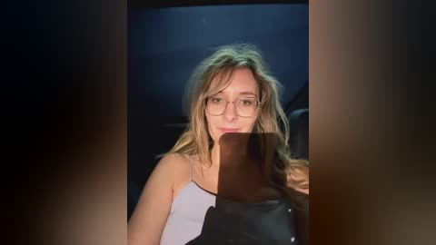 Media: Video of a young woman with messy blonde hair, wearing glasses, and a black and white striped top, seated in a dark car, looking slightly to the side.