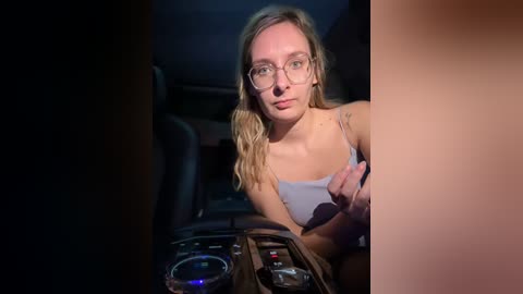 Media: Video of a blonde woman with glasses, wearing a light gray tank top, sitting in a dimly lit car with a center console and cup holder visible.