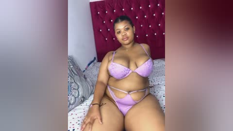 Media: Video of a plus-sized Black woman with medium brown skin, wearing a lavender lace lingerie set, sitting on a bed with white and grey patterned bedding and a tufted red headboard.