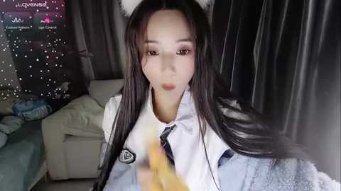 Media: Video of an East Asian woman with long black hair, wearing a white blouse, tie, and cat ears, sitting in a dimly lit room with dark curtains, holding a yellow banana.