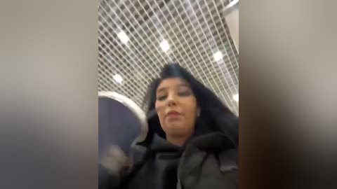 Media: Video of a person with a dark complexion and short hair, wearing a black jacket, standing in a tiled room with a grid-patterned ceiling. The image is slightly blurry.